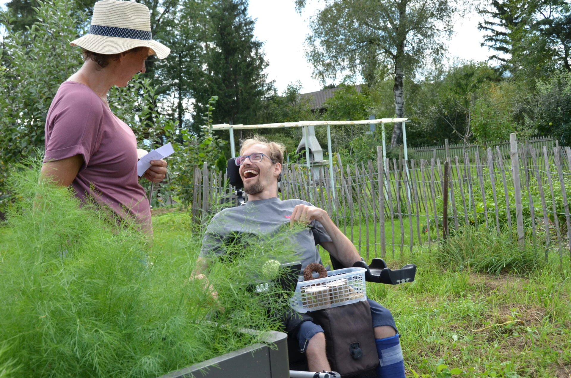 Mann im Rollstuhl und Betreuerin lachen gemeinsam im Schrebergarten zwischen Kraeuterbeeten