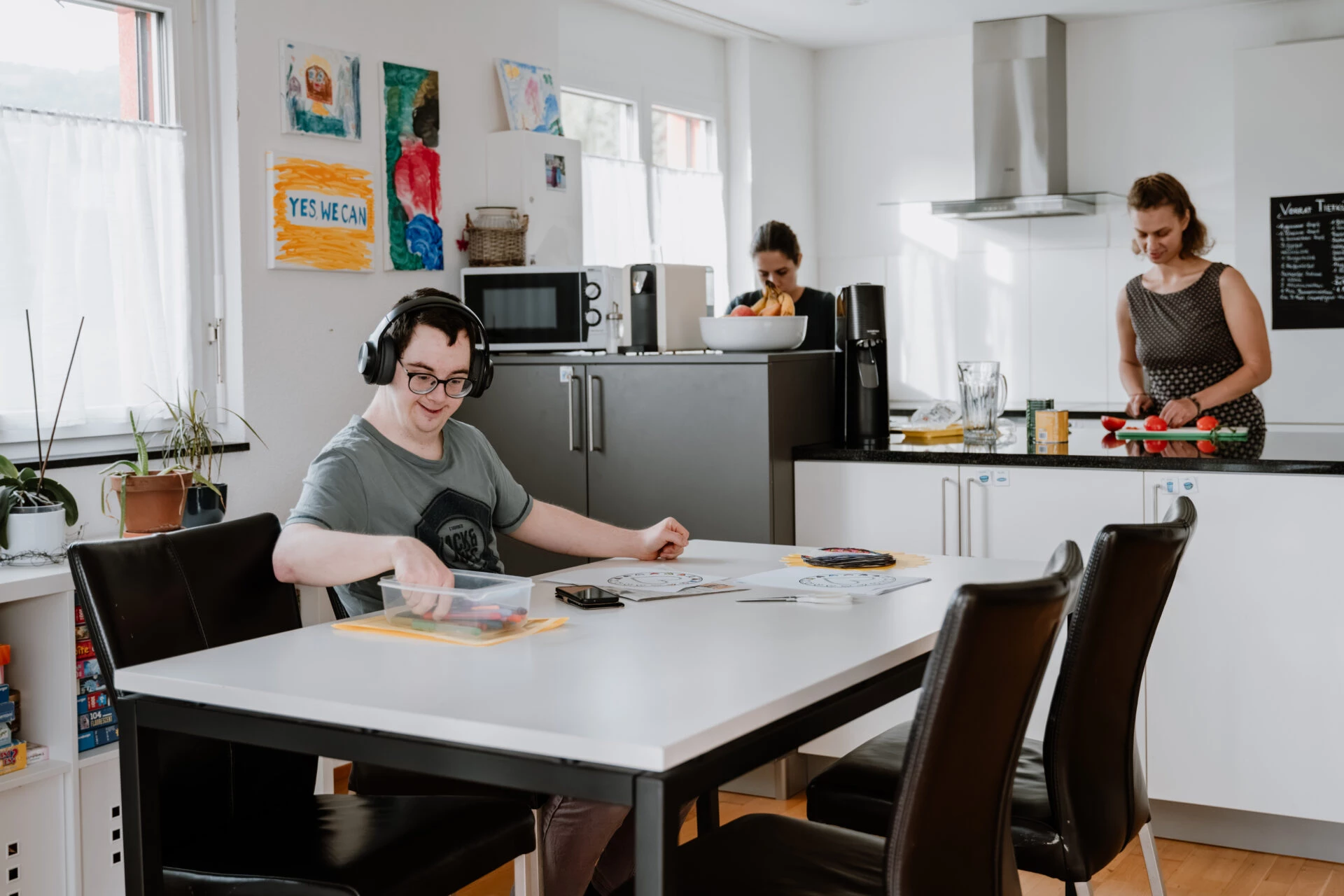 Bewohner mit Kopfhoerern am Esstisch, im Hintergrund bereiten zwei Frauen das Essen in der Kueche vor