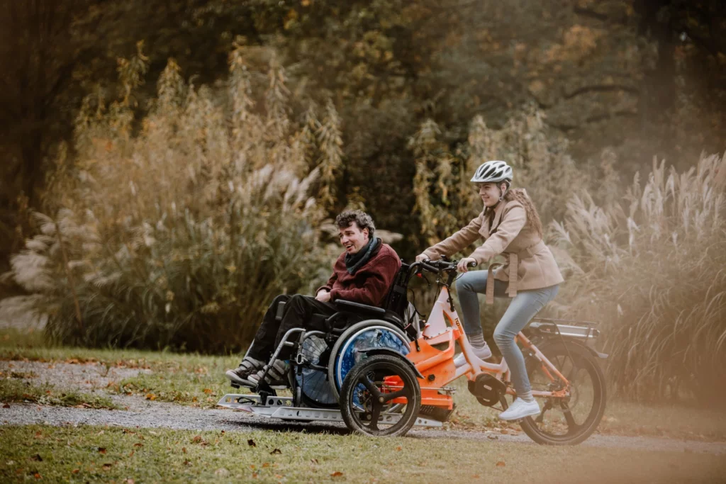 Mann im Rollstuhl wird von einer Begleiterin mit einem Rollstuhl-Velo durch einen herbstlichen Park gefahren