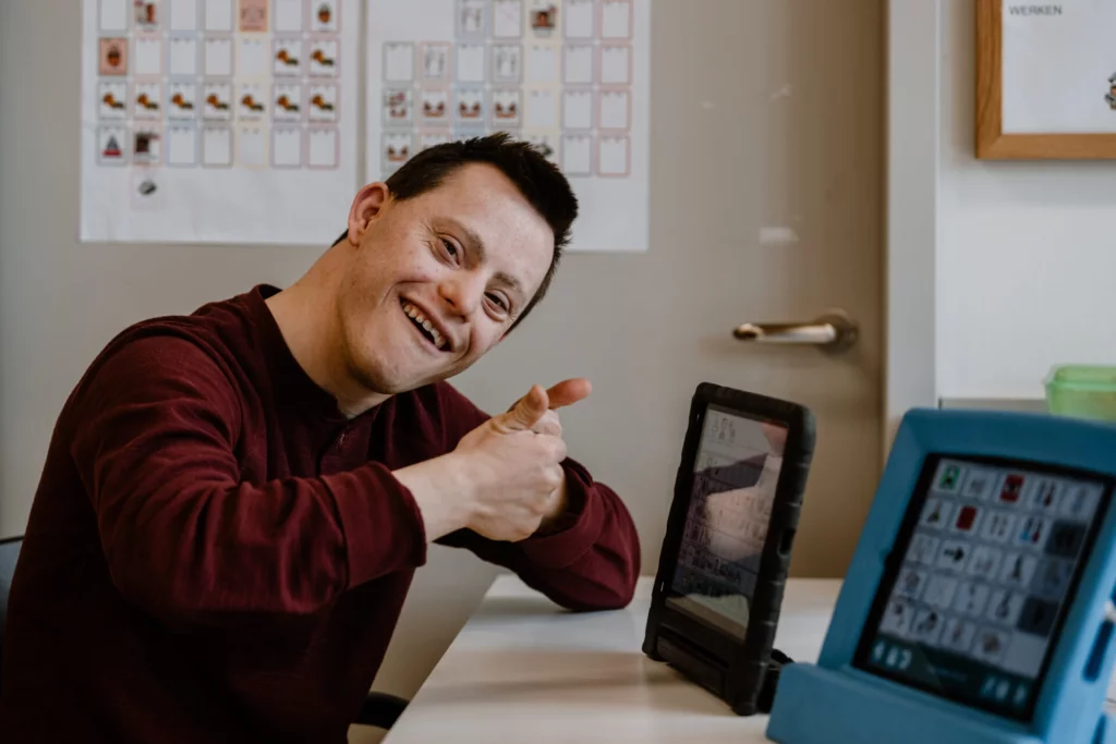 Lachender junger Mann zeigt auf ein Tablet mit Kommunikationshilfe am Arbeitsplatz