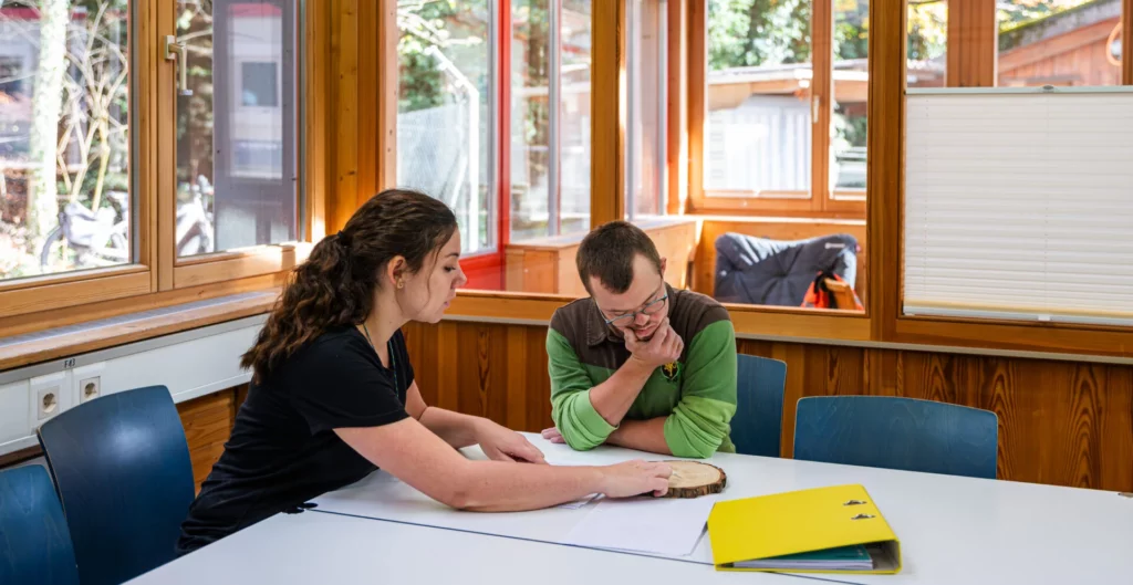 Betreuerin und Bewohner schauen gemeinsam Unterlagen an einem Tisch im Besprechungsraum an