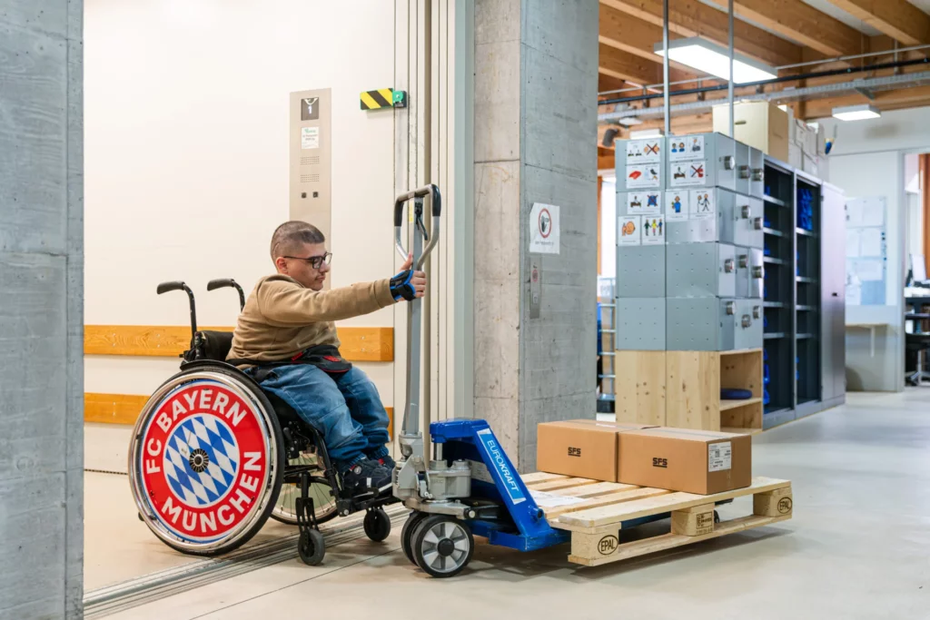 Mann im Rollstuhl transportiert Pakete auf einer Palette mit einem Hubwagen in der Werkstatt