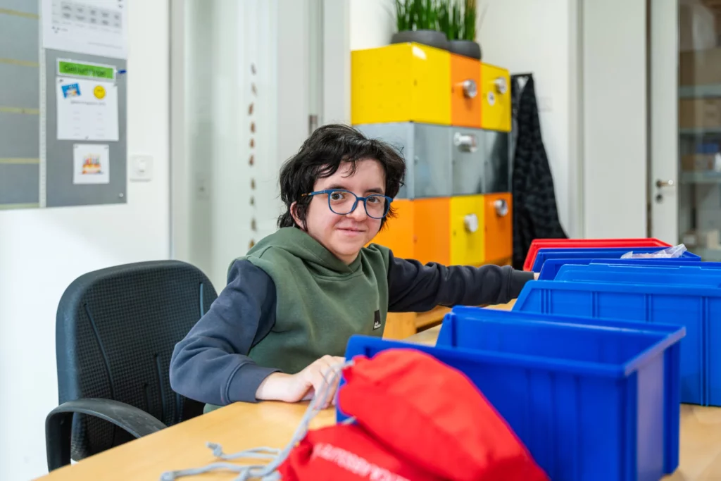 Junge Frau mit Brille sortiert lachend farbige Kunststoffboxen am Arbeitsplatz