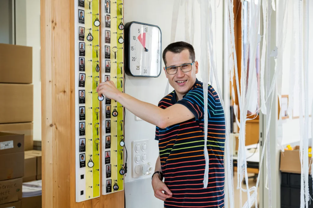 Lachender Mann mit Brille steht an einer Stempeluhr mit Fototafel in der Werkstatt