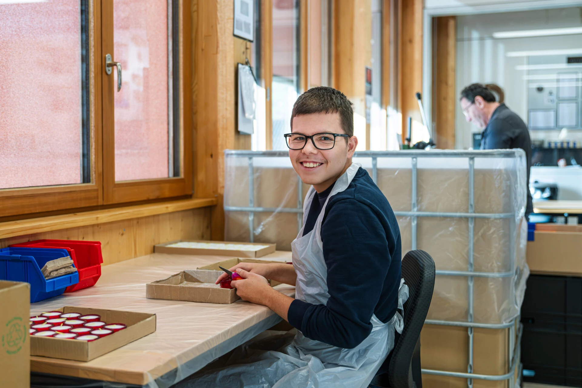 Junger Mann mit Brille und Schuerze sitzt lachend am Arbeitsplatz und verpackt Produkte in Kartons