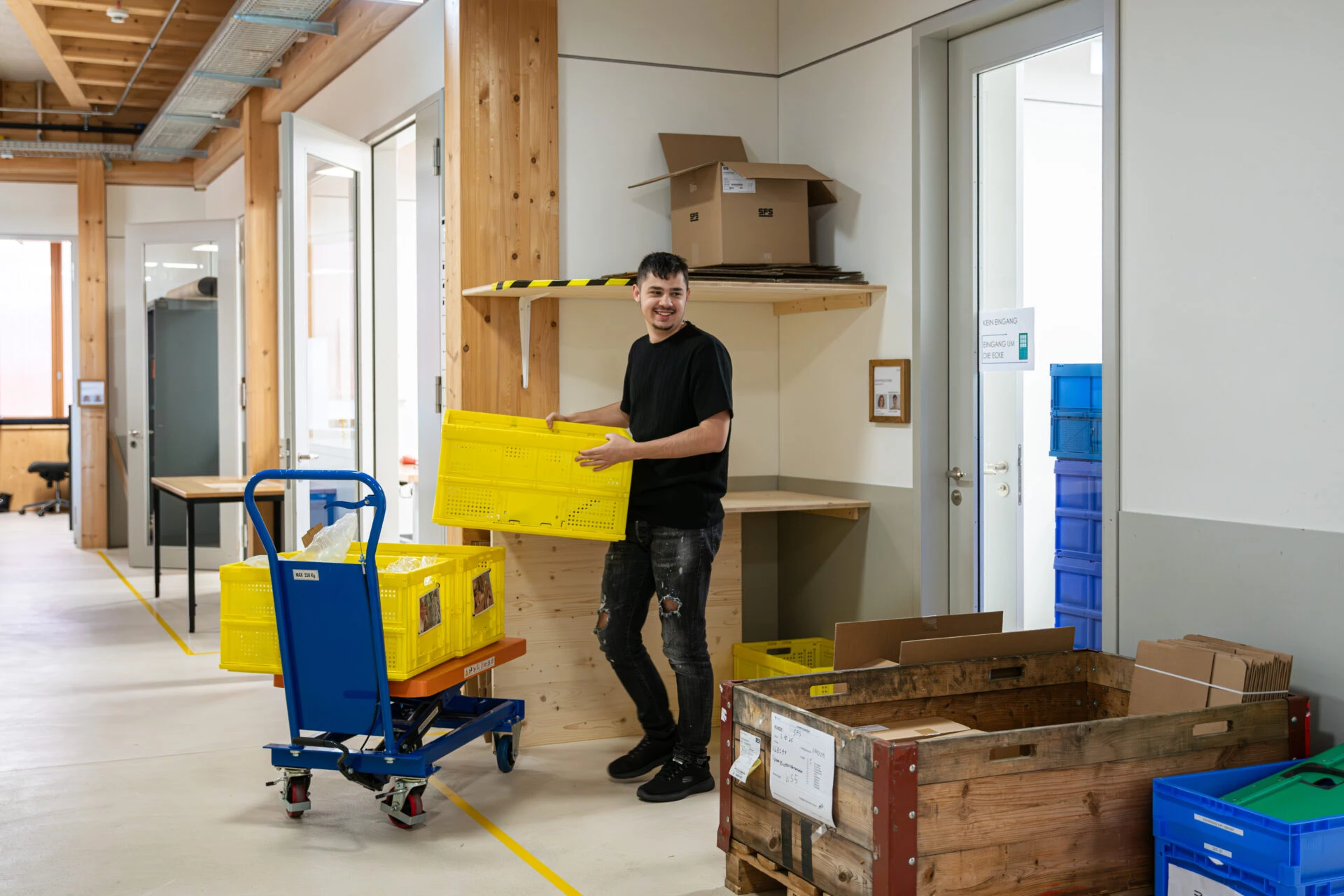 Mann traegt eine gelbe Transportkiste in einem hellen Lager- und Logistikbereich mit Rollwagen