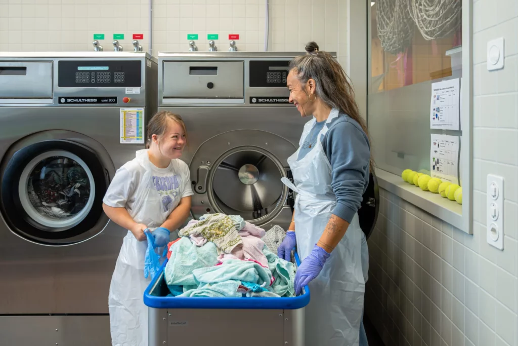 Zwei Frauen stehen lachend vor Industriewaschmaschinen in der Waescherei und halten einen Waeschekorb
