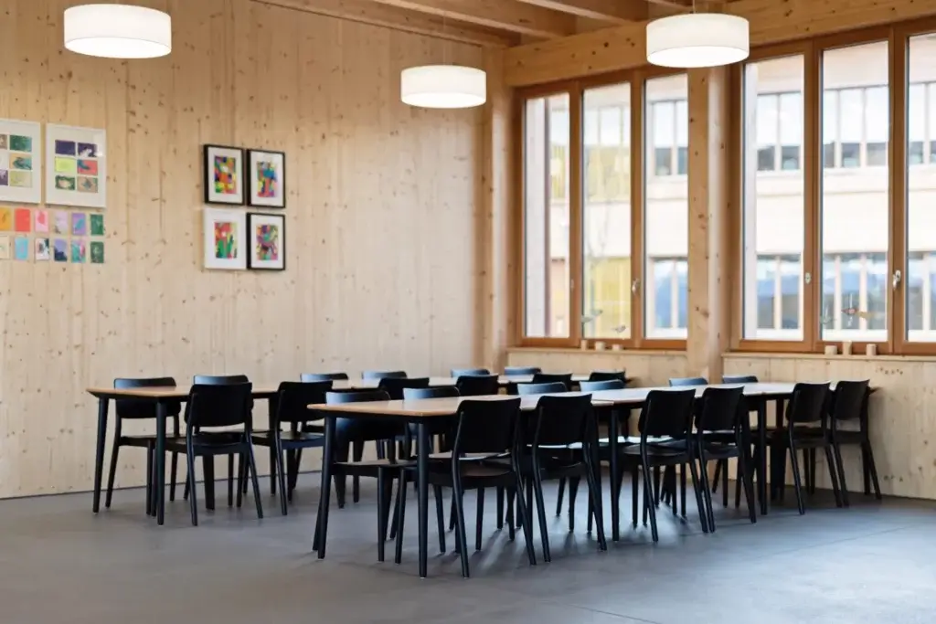 Gemeinschaftsraum mit Tischen, Stuehlen und farbigen Kunstwerken an der Holzwand