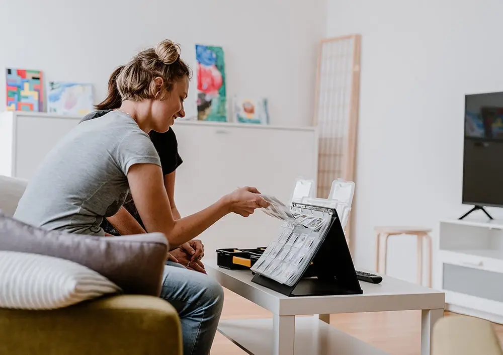 Bewohnerin sortiert lachend Medikamentenbox im hellen Wohnzimmer mit bunten Bildern an der Wand