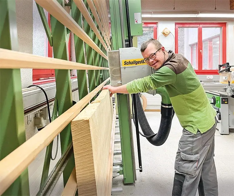 Mitarbeiter bedient laechelnd eine Plattensaege in der Holzwerkstatt von Rhyboot