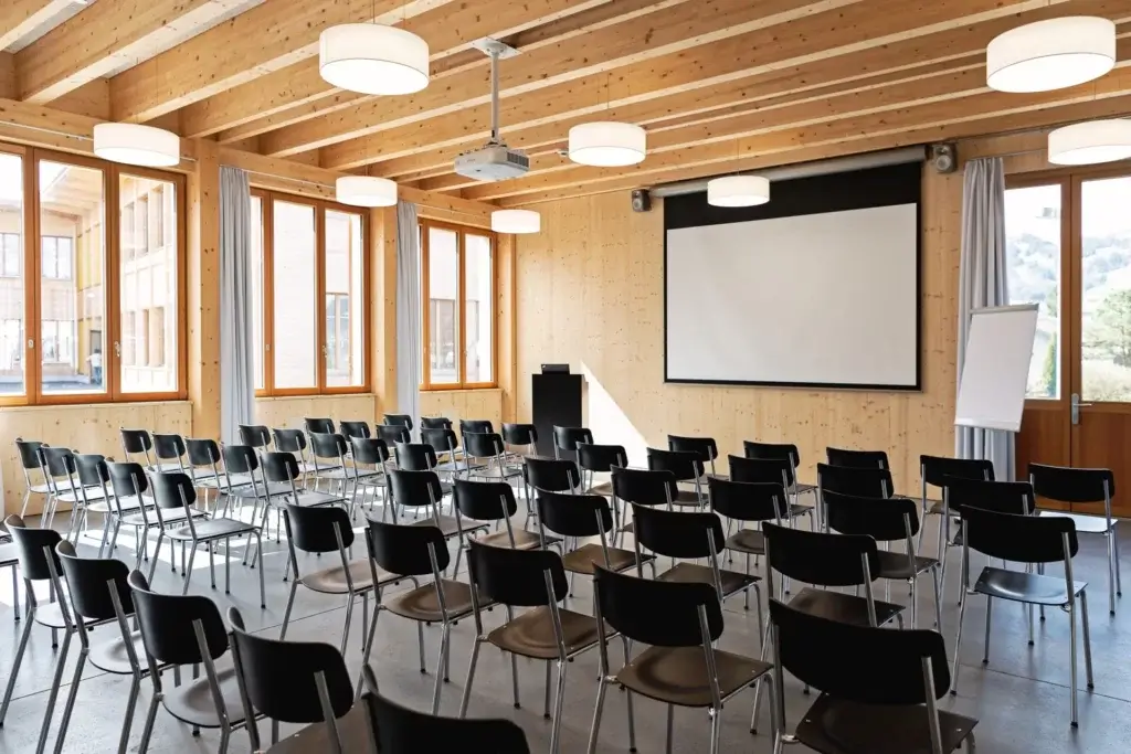 Heller Mehrzweckraum in Holzbauweise mit Stuhlreihen, Leinwand und Beamer fuer Veranstaltungen