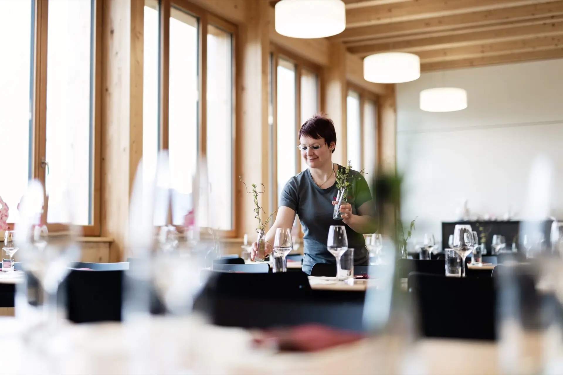Mitarbeiterin dekoriert Tische mit Blumen im lichtdurchfluteten Restaurant mit Holzverkleidung