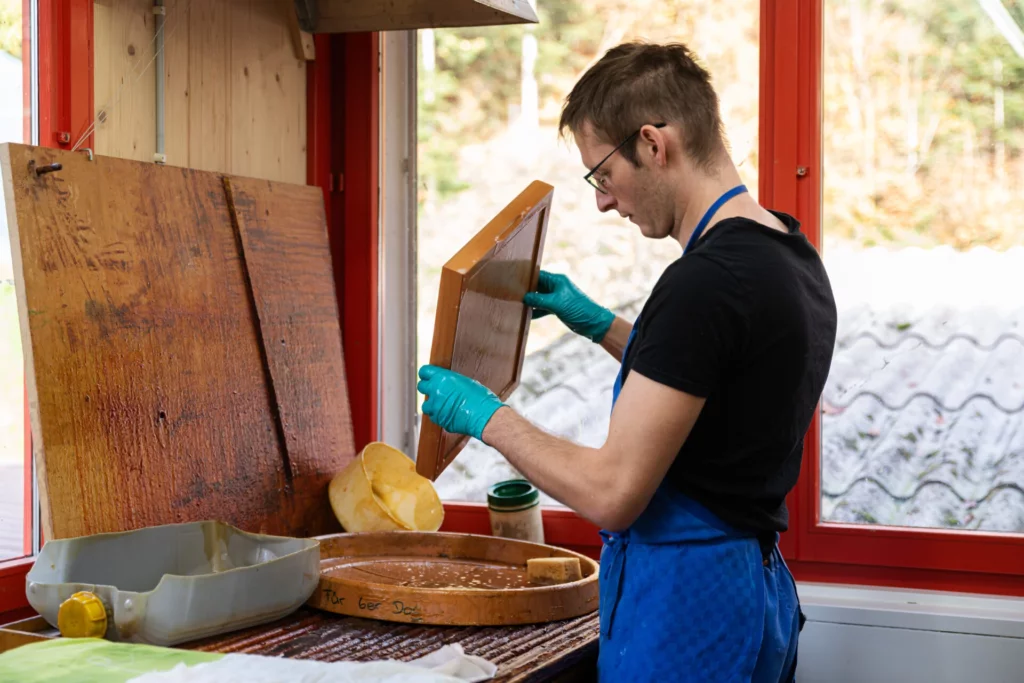 Mitarbeiter in Schuerze hebt handgeschoepftes Papier aus einem Wasserbecken in der Werkstatt