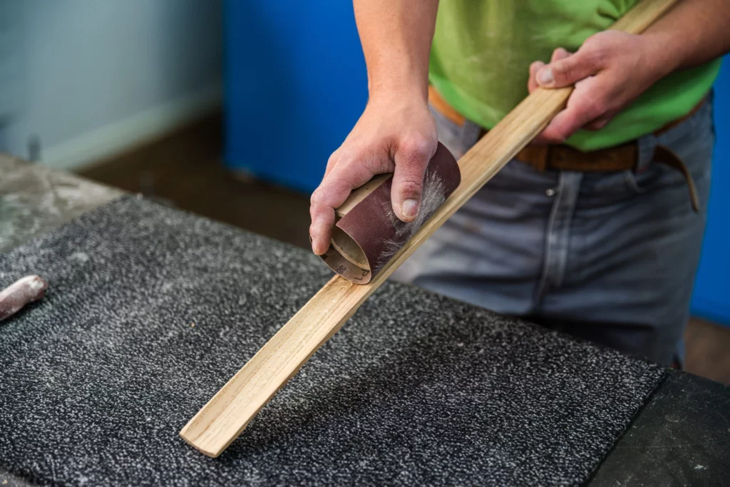 Nahaufnahme von Haenden beim Schleifen einer Holzleiste mit Schleifpapier
