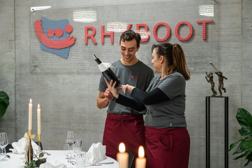 Mitarbeiter und Betreuerin betrachten lachend eine Weinflasche vor der Rhyboot-Logowand