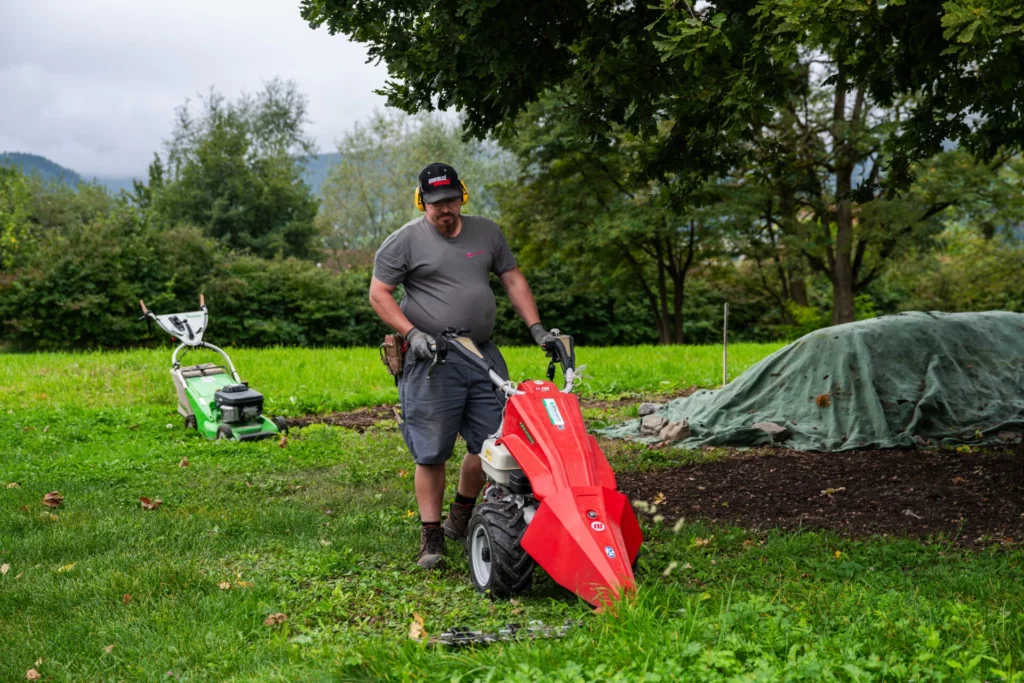 Mitarbeiter bedient eine rote Motorhacke auf einer gruenen Wiese bei der Gartenarbeit