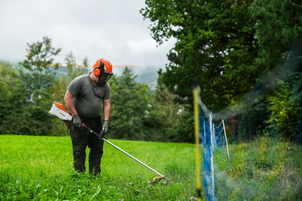 Mitarbeiter mit Schutzhelm und Gehoerschutz maeht Gras mit einer Motorsense
