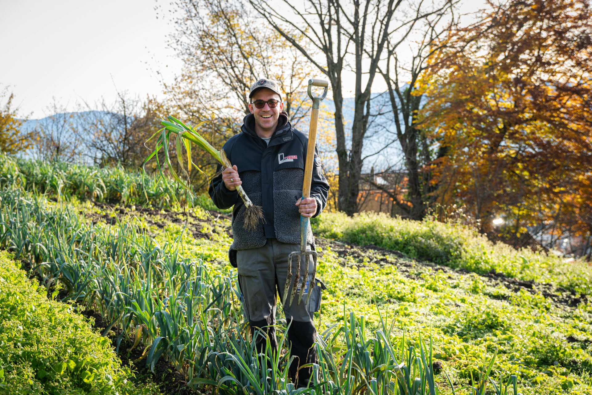 Mitarbeiter steht mit Spaten und frisch geerntetem Lauch auf dem herbstlichen Feld
