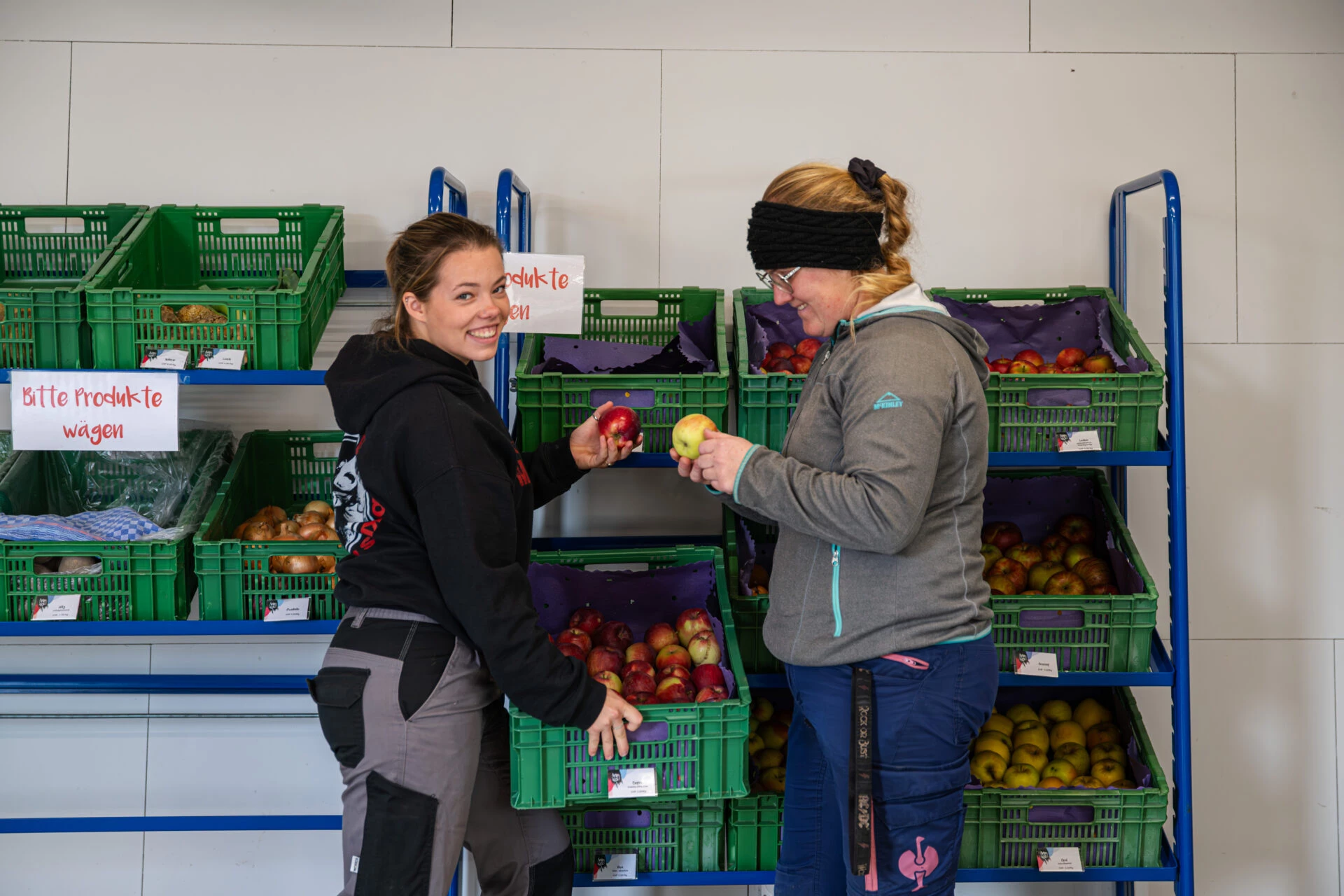 Zwei Frauen zeigen sich gegenseitig Aepfel vor Regalen voller frischem Obst und Gemuese