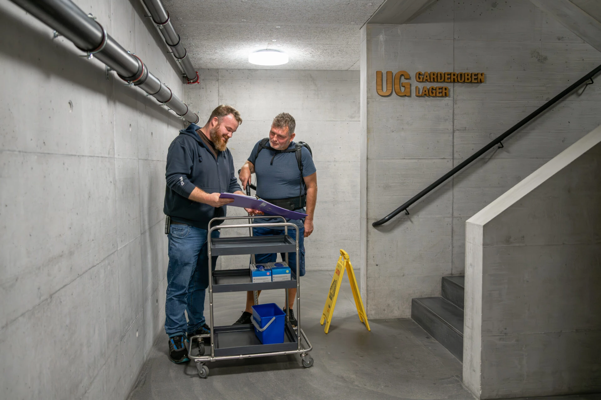 Zwei Mitarbeiter besprechen einen Reinigungsplan im Untergeschoss neben einem Reinigungswagen