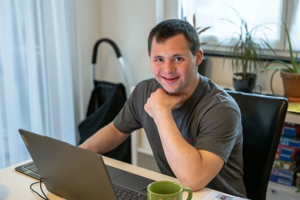 Laechelnder junger Mann sitzt mit Laptop und Kaffeetasse am Schreibtisch im Wohnbereich